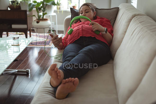Joven embarazada acostada en el sofá mujer colocando auriculares en su vientre en casa - foto de stock