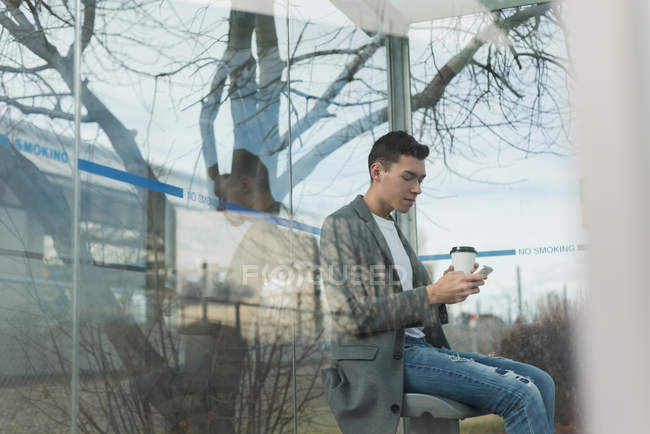 Homem usando telefone celular enquanto toma café na parada de ônibus — Fotografia de Stock