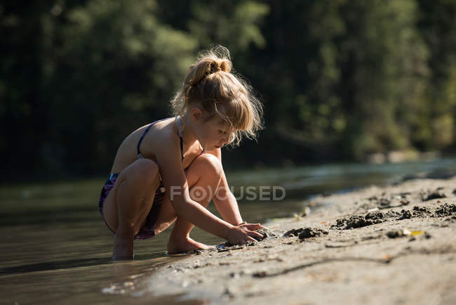 Menina brincando com areia perto da margem do rio em um dia ensolarado — Fotografia de Stock