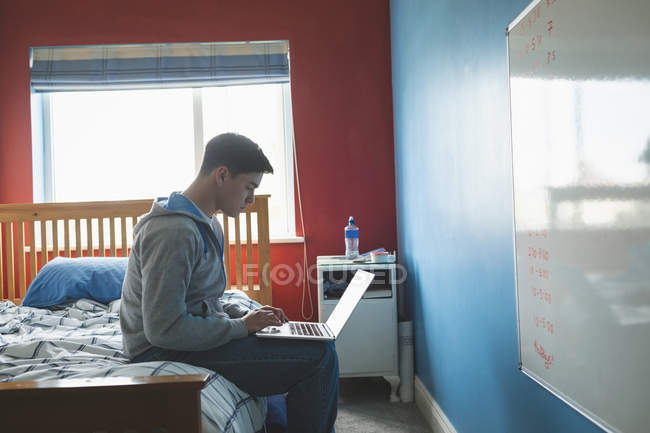 Junger Mann mit Laptop im Schlafzimmer auf dem Bett sitzend, Seitenansicht. — Stockfoto