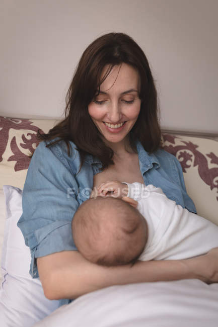 Sorrindo jovem mãe sentada na cama amamentando seu bebê em casa — Fotografia de Stock