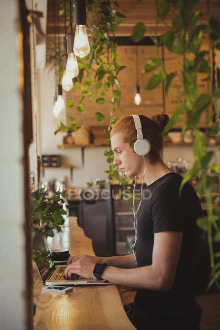 Hombre escuchando música en los auriculares mientras usa el ordenador portátil en la cafetería - foto de stock