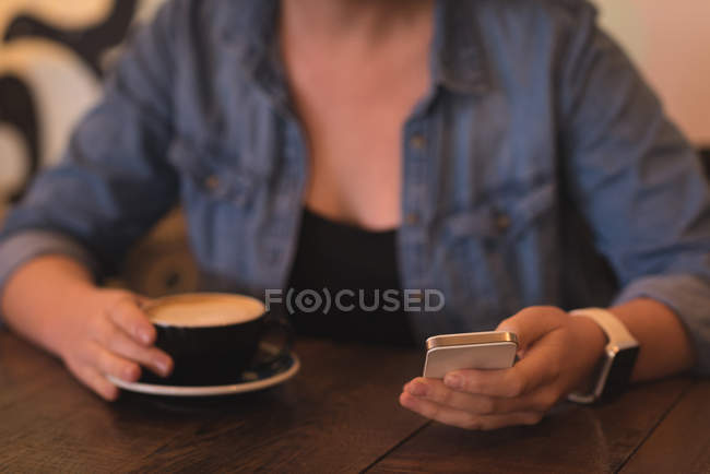 Sección media de la mujer que usa el teléfono móvil mientras toma café en la cafetería - foto de stock