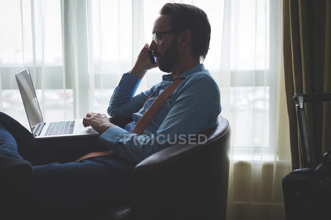 Empresário falando no celular enquanto usa laptop no quarto de hotel — Fotografia de Stock
