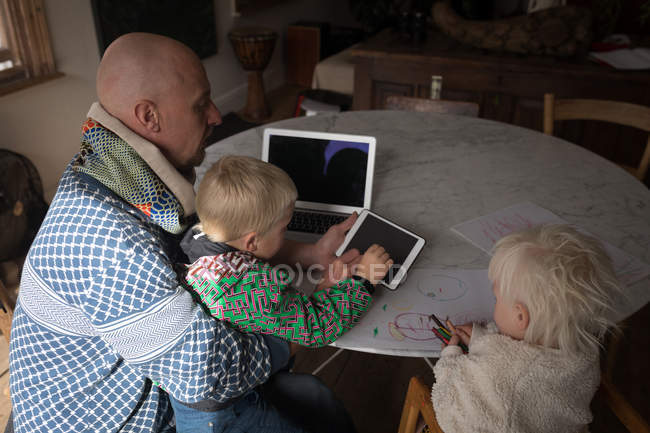 Father and kids using digital tablet and laptop at table in living room at home. — Stock Photo