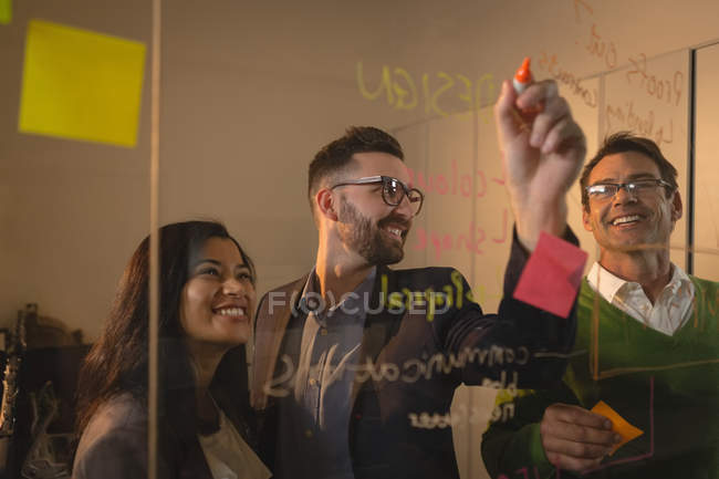 Gente de negocios sonriente discutiendo ideas en la pared de vidrio y notas adhesivas en la oficina . - foto de stock