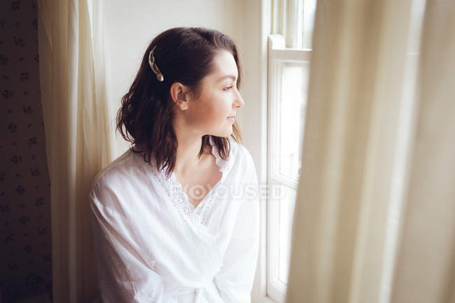 Belle femme regardant par la fenêtre par une journée ensoleillée — Photo de stock