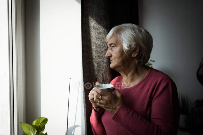 Mujer mayor reflexiva tomando una taza de té mientras mira por la ventana - foto de stock