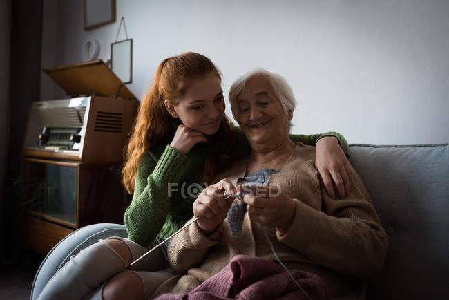 Neta sorridente olhando para a avó enquanto tricotava lã em casa — Fotografia de Stock