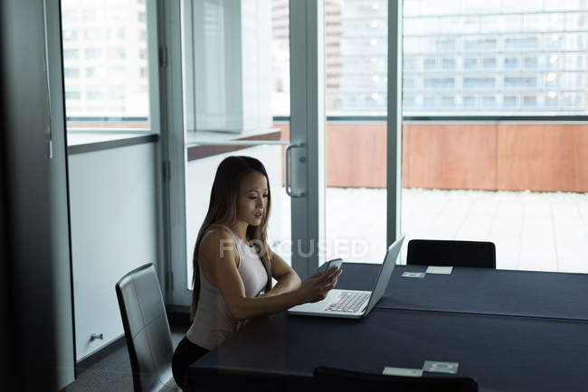 Asiática mujer de negocios sentado solo trabajando en portátil usando su móvil en la oficina - foto de stock