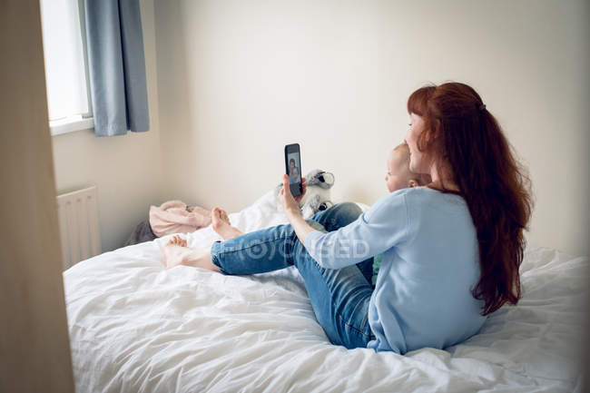 Mother and baby girl taking selfie with mobile phone in bedroom at home — Stock Photo