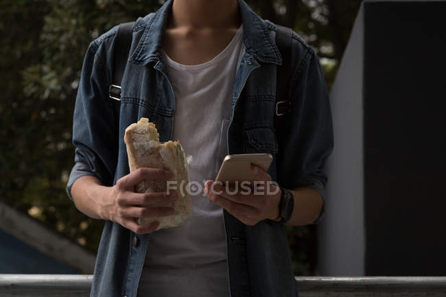 Молодий чоловік використовує мобільний телефон, маючи бургер в сходах — стокове фото