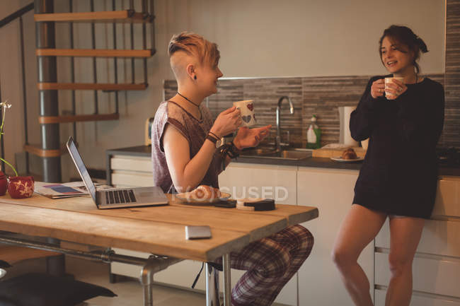 Mujeres jóvenes hablando con tazas de café en la cocina en casa . - foto de stock