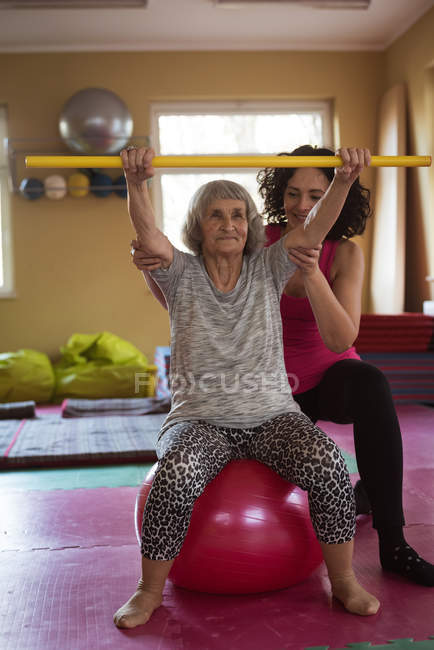 Женщина-терапевт помогает пожилой женщине с палкой для упражнений и мячом для упражнений в доме престарелых — стоковое фото