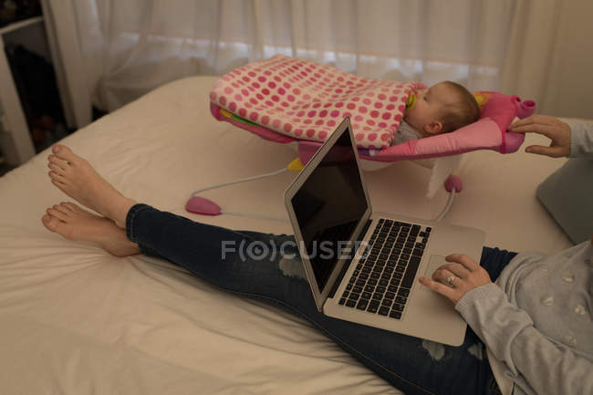 Madre utilizzando il computer portatile mentre il bambino dorme sul letto a casa . — Foto stock