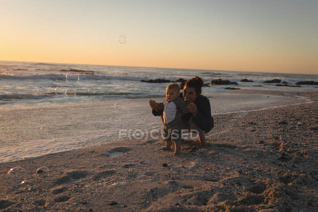 Mère et fils soufflant la bulle avec la baguette à bulles à la plage pendant le coucher du soleil — Photo de stock