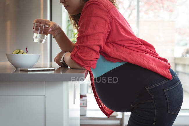 Sección media de la mujer embarazada bebiendo agua en la cocina - foto de stock
