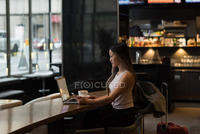 Geschäftsfrau hält Handy in der Hand, während sie in der Cafeteria am Laptop arbeitet — Stockfoto