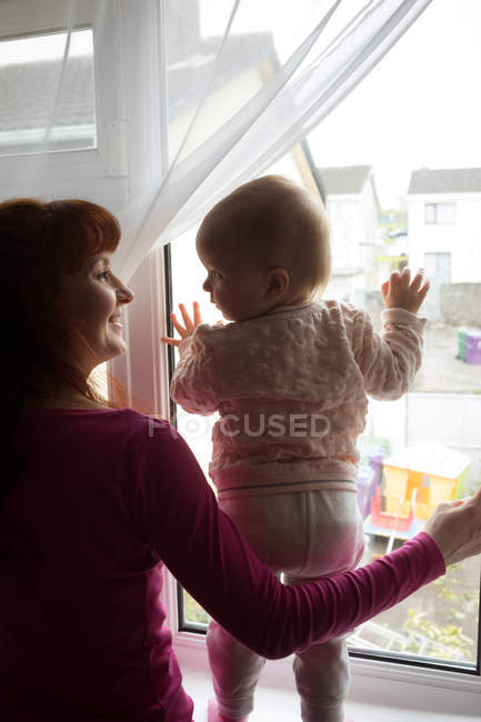Mother with her baby girl looking through window at home — Stock Photo