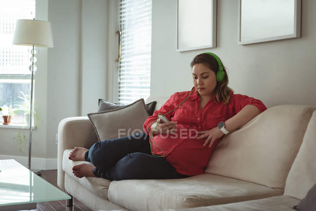 Young pregnant woman sitting on sofa listing to music on her mobile phone at home — Stock Photo