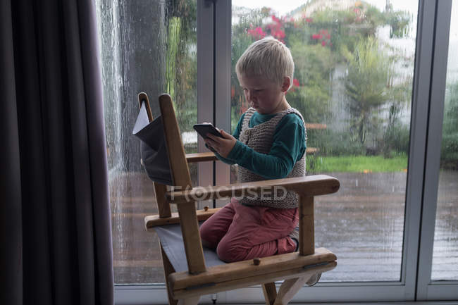 Хлопчик використовує мобільний телефон, сидячи на стільці вдома . — стокове фото