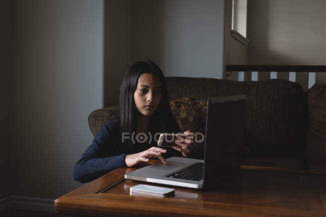 Девочка пользуется ноутбуком и мобильным телефоном в гостиной дома — стоковое фото