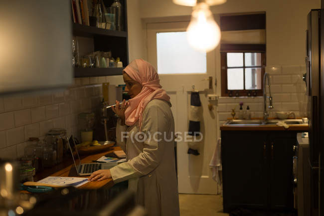Мусульманка разговаривает по телефону и пользуется ноутбуком на кухне — стоковое фото