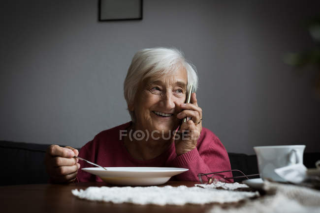 Mujer mayor sonriente hablando por teléfono móvil mientras desayuna en la sala de estar - foto de stock