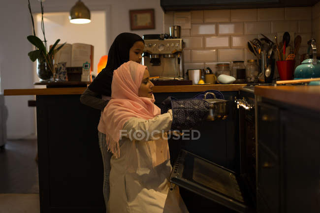 Mujer musulmana e hija usando el horno en la cocina en casa - foto de stock
