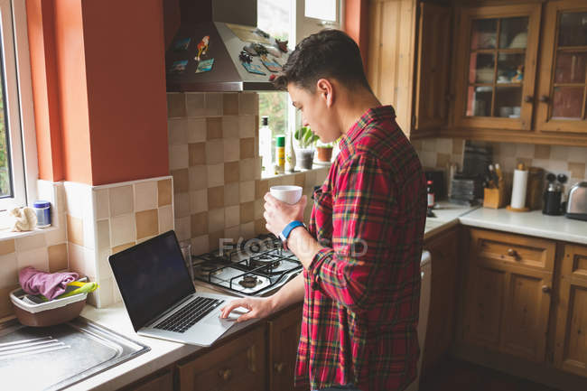 Чоловік має каву під час використання ноутбука на кухні вдома . — стокове фото