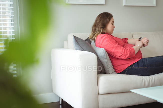 Mulher grávida sentada no sofá verificando seu relógio inteligente em casa — Fotografia de Stock