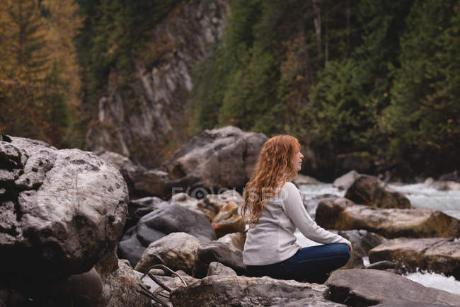 Visão traseira da mulher sentada nas rochas perto de um rio fluindo — Fotografia de Stock