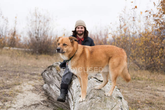 Uomo sorridente e il suo cane seduto su tronco di legno — Foto stock