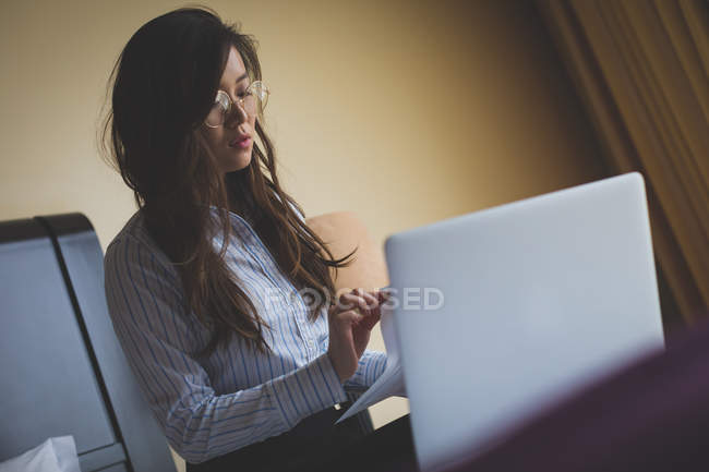 Femme d'affaires détenant des documents tout en travaillant sur ordinateur portable dans la chambre d'hôtel — Photo de stock