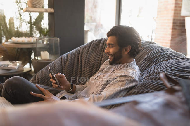 Hombre usando el teléfono móvil mientras se relaja en el sillón en la cafetería - foto de stock