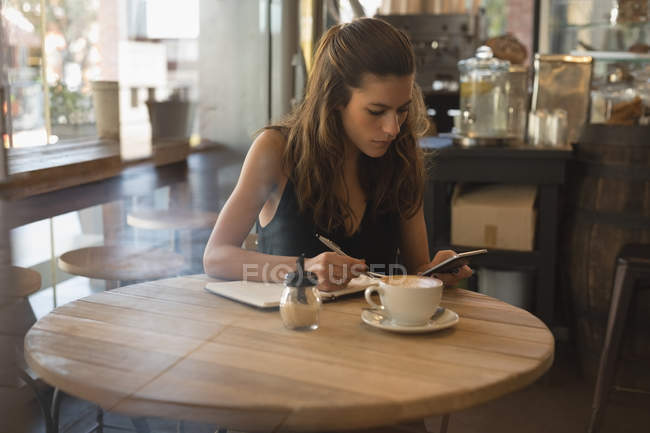 Жінка використовує мобільний телефон під час написання на щоденнику в кав'ярні — стокове фото