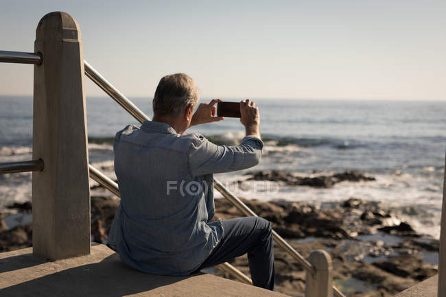 Вид ззаду старшого чоловіка, який фотографує з мобільного телефону біля морської сторони — стокове фото