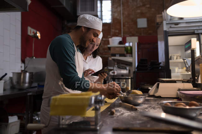 Baker preparando pasta mientras compañero de trabajo usando tableta digital su lado en la panadería - foto de stock