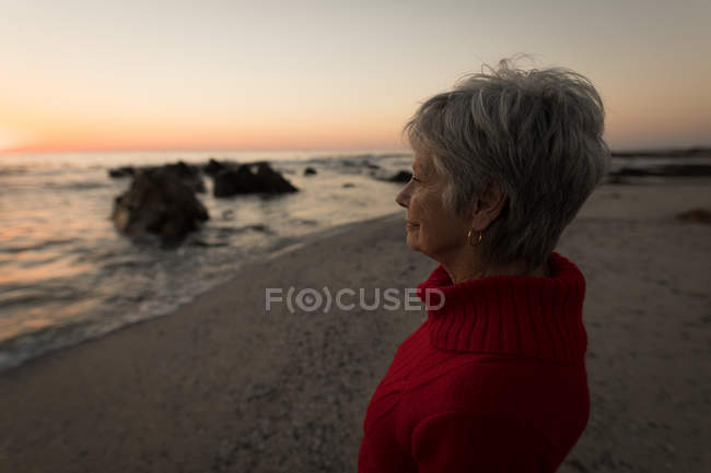 Mulher sênior pensativo em pé na praia durante o pôr do sol — Fotografia de Stock