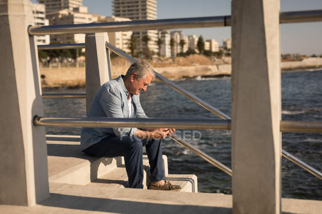 Hombre mayor usando el móvil cerca del mar en el paseo marítimo - foto de stock