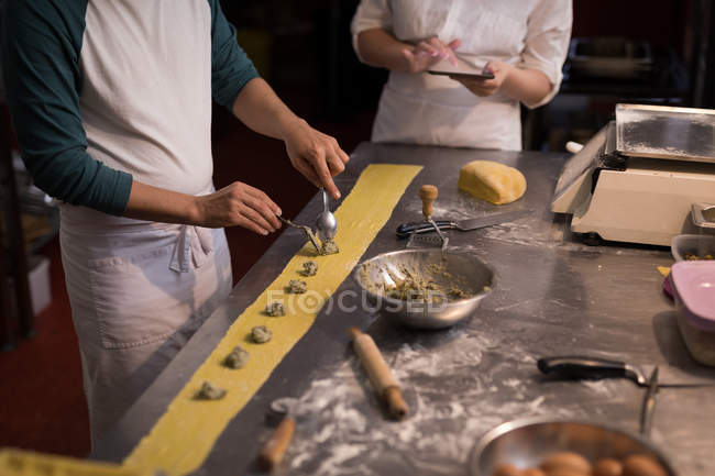 Baker preparando pasta mientras compañero de trabajo usando tableta digital su lado en la panadería - foto de stock