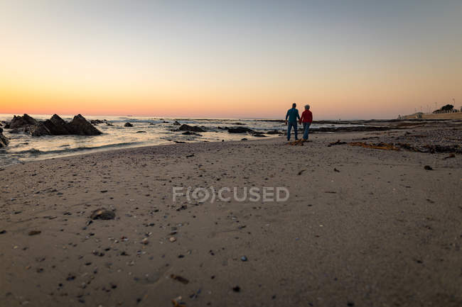 Vista trasera de pareja mayor caminando en la playa al atardecer - foto de stock