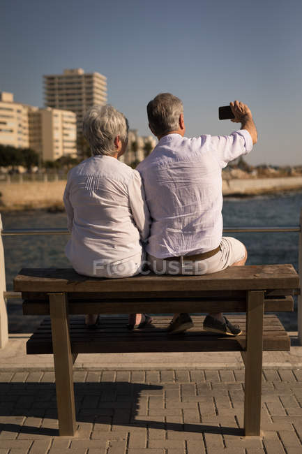 Вид ззаду старшої пари, що приймає селфі, сидячи на лавці на набережній — стокове фото