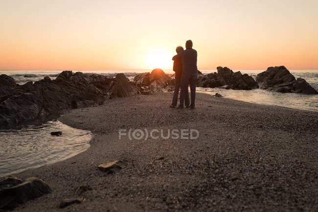 Visão traseira do casal sênior abraçando na praia durante o pôr do sol — Fotografia de Stock