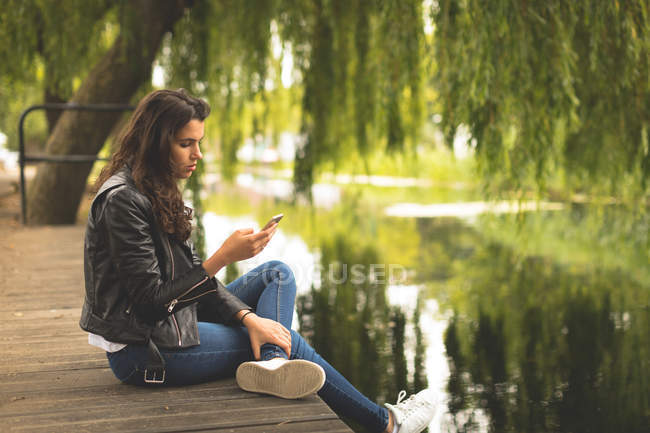 Hermosa mujer usando el teléfono móvil mientras está sentado cerca del lago - foto de stock