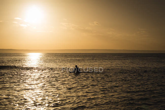 Surfista con tabla de surf surfeando en el mar al atardecer - foto de stock