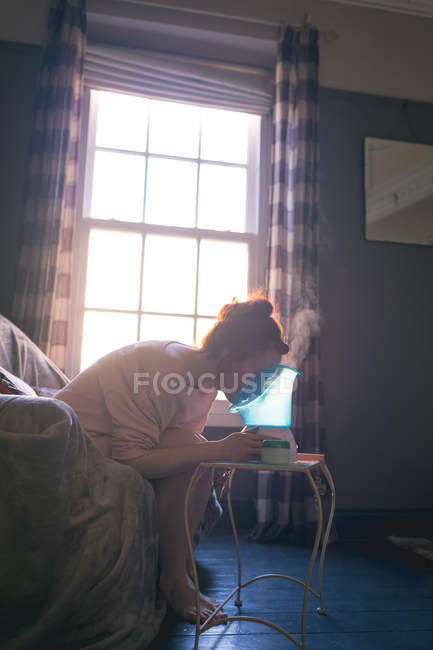 Mujer usando vaporizador facial en sofá en casa - foto de stock