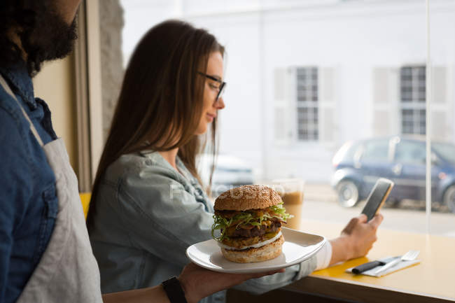 Camarero sirviendo hamburguesa a la mujer mientras usa el teléfono móvil en la cafetería - foto de stock