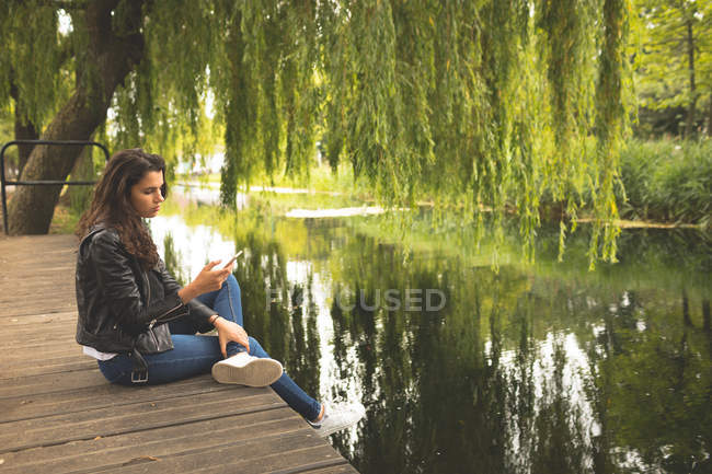 Mulher bonita usando telefone celular enquanto sentado perto do lado do lago — Fotografia de Stock