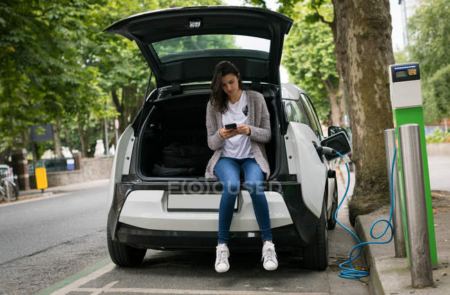 Женщина с мобильного телефона во время зарядки электромобиля на зарядной станции — стоковое фото
